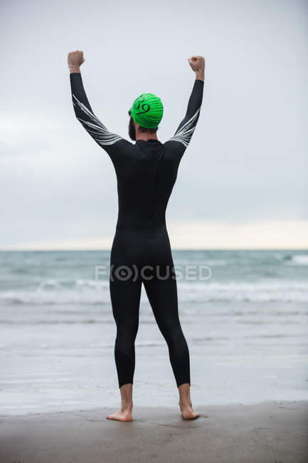 Rear view of happy athlete standing on beach with hands raised — Stock Photo