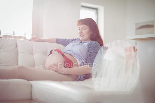 Вагітна жінка з навушниками на животі розслабляється на дивані у вітальні — стокове фото