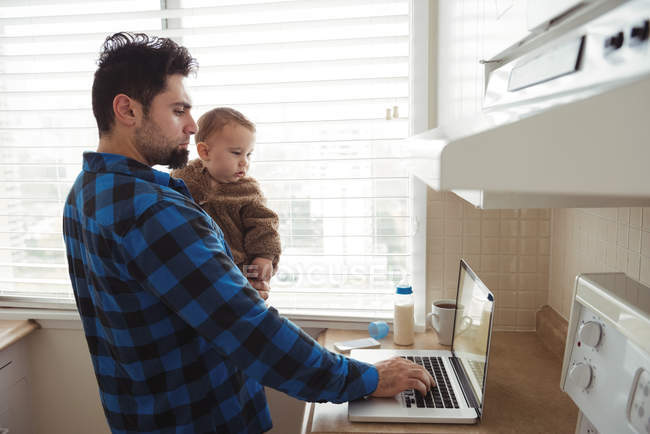 Padre che usa il computer portatile mentre tiene il bambino in cucina a casa — Foto stock