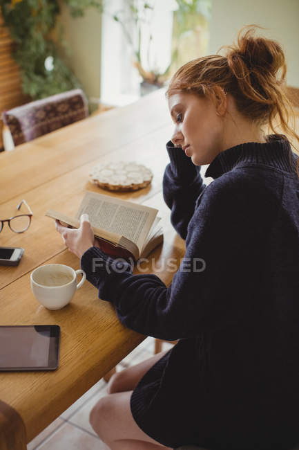 Femme assise à table et lisant un livre à la maison — Photo de stock