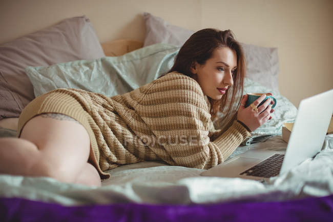 Belle femme utilisant un ordinateur portable tout en prenant un café sur le lit à la maison — Photo de stock
