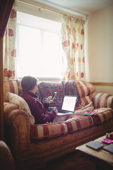 Hermosa mujer sosteniendo la taza de café mientras usa el ordenador portátil y el teléfono móvil en el sofá en casa - foto de stock