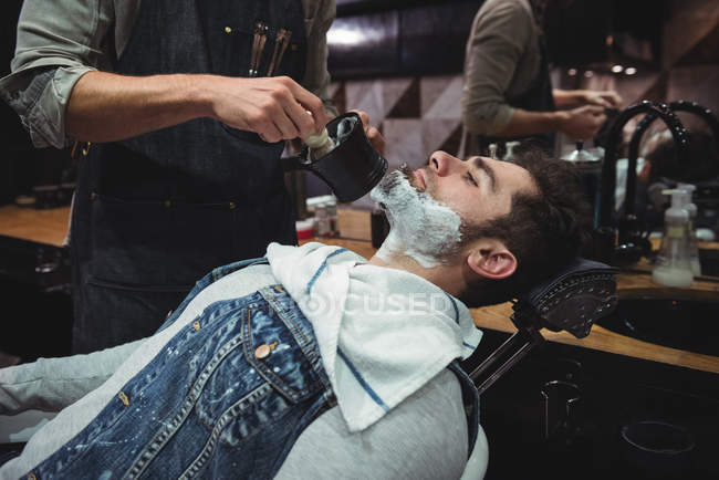 Sección media del peluquero aplicando crema en la barba del cliente en la peluquería - foto de stock