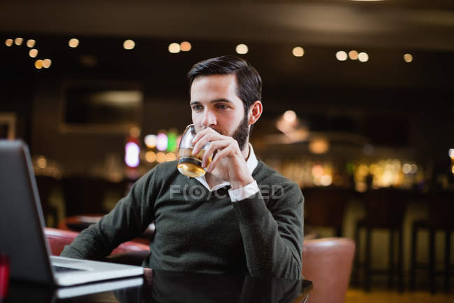 Чоловік дивиться на ноутбук, маючи келих напою в барі — стокове фото