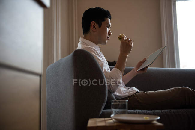 Homem usando tablet digital enquanto tem sanduíche em casa — Fotografia de Stock