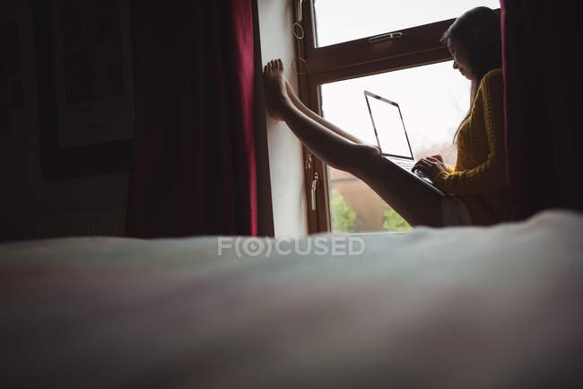 Femme utilisant un ordinateur portable tout en étant assis au rebord de la fenêtre à la maison — Photo de stock