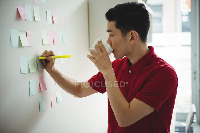 Ejecutivo de negocios escribiendo en notas adhesivas mientras toma una taza de café en la oficina - foto de stock