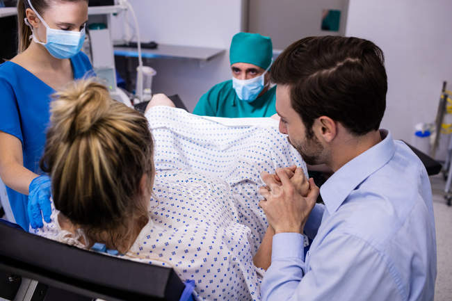 Squadra medica che esamina la donna incinta durante il parto mentre l'uomo le tiene la mano in sala operatoria — Foto stock