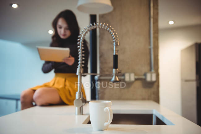 Женщина сидит на кухонном столе с помощью цифрового планшета дома — стоковое фото