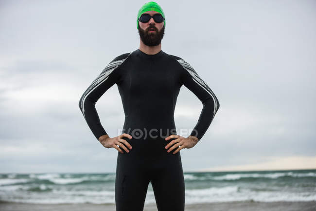 Retrato de atleta en traje mojado de pie con las manos en la cintura en la playa - foto de stock