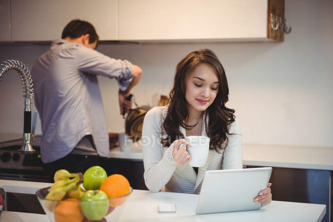 Frau nutzt digitales Tablet, während Mann im Hintergrund in der Küche arbeitet — Stockfoto