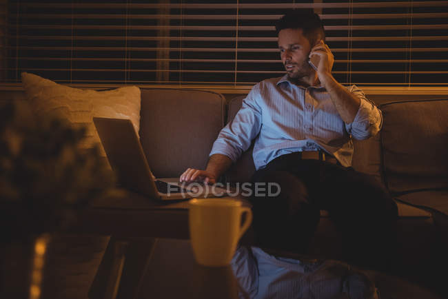Homme parlant sur un téléphone portable tout en utilisant un ordinateur portable dans le salon à la maison — Photo de stock