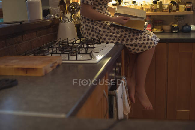 Frau sitzt auf Küchenarbeitsplatte und liest Buch zu Hause — Stockfoto