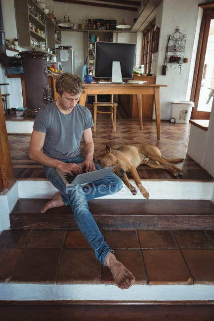 Hombre sentado en el suelo y usando portátil en casa, perro acostado a su lado - foto de stock