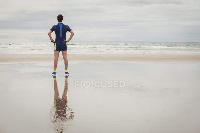Вид сзади спортсмена, стоящего с руками на бедрах на пляже — стоковое фото