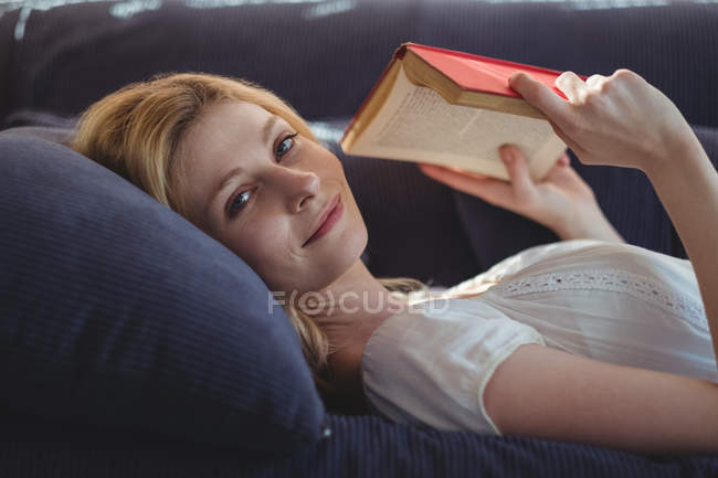Retrato de mulher bonita deitada no sofá e livro de leitura na sala de estar em casa — Fotografia de Stock