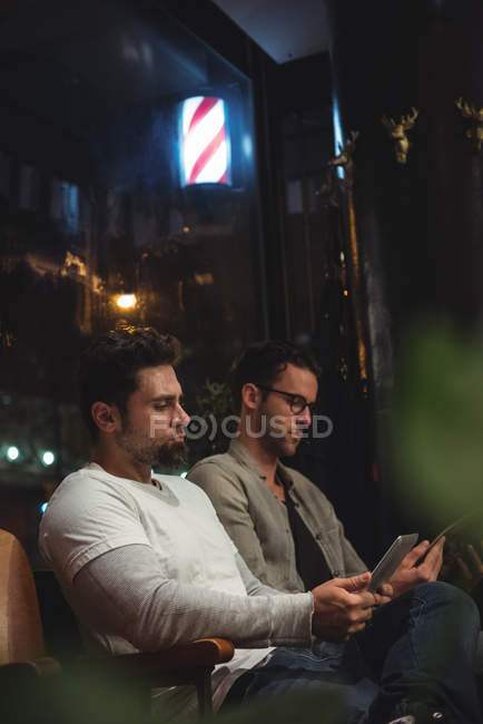 Клиенты, использующие цифровой планшет и мобильный телефон во время ожидания в парикмахерской — стоковое фото