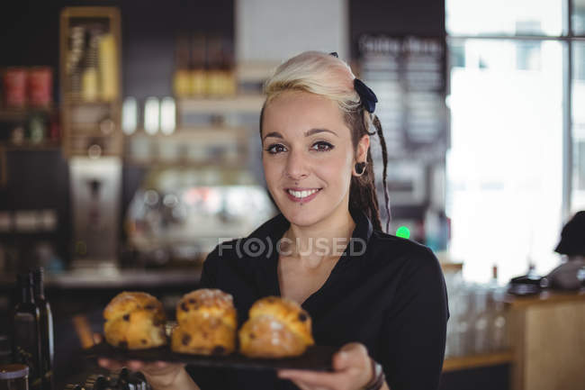 Retrato de camarera sosteniendo bandeja de magdalenas en el mostrador en la cafetería - foto de stock