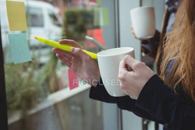 Ejecutivo de negocios escribiendo en notas adhesivas mientras toma una taza de café en la oficina - foto de stock