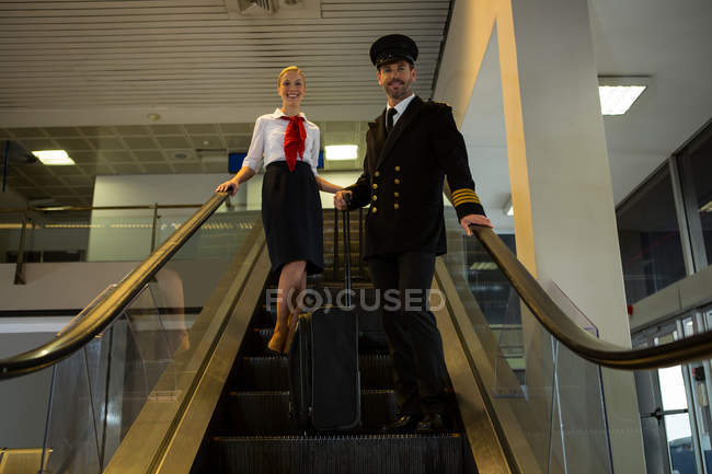 Пилот и стюардесса со своими тележками стоят на эскалаторе в терминале аэропорта — стоковое фото
