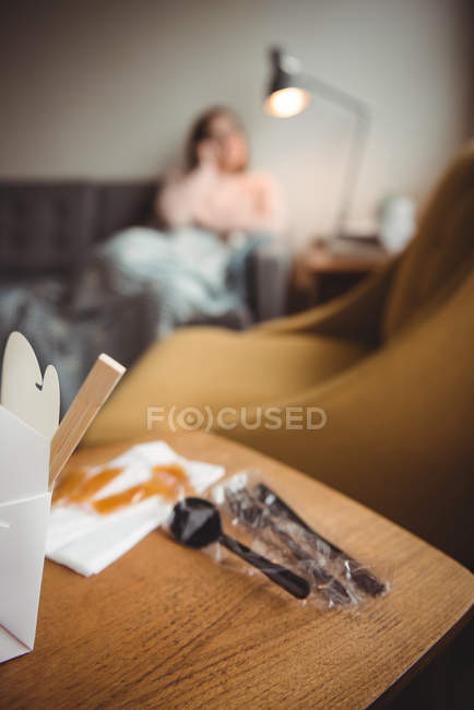 Caja de comida y cucharas en la mesa mientras la mujer se relaja en el fondo en casa - foto de stock