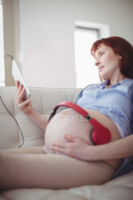 Schwangere mit Kopfhörern auf dem Bauch entspannt sich auf Sofa im Wohnzimmer — Stockfoto