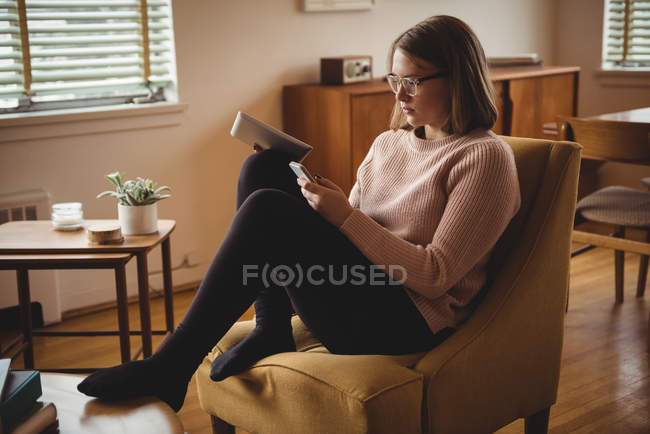 Donna seduta sulla sedia con cellulare e tablet digitale in salotto — Foto stock