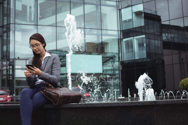 Mujer de negocios sentada cerca de la fuente y usando teléfono móvil fuera del edificio de oficinas - foto de stock