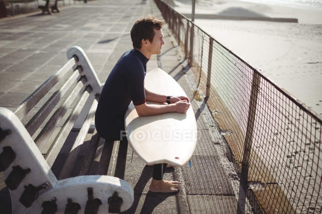 Thoughtful surfer sitting with surfboard on pier at beach — Stock Photo