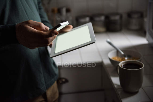 Средняя часть человека с помощью цифрового планшета и мобильного телефона на кухне в домашних условиях — стоковое фото