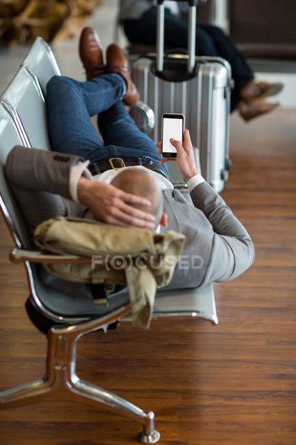 Businessman using mobile phone while lying on chairs in waiting area at airport terminal — Stock Photo