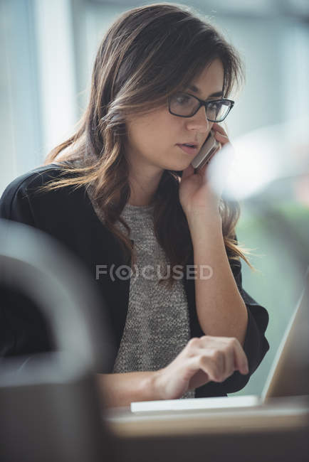 Ejecutivo de negocios hablando en el teléfono móvil mientras usa el ordenador portátil en la oficina - foto de stock