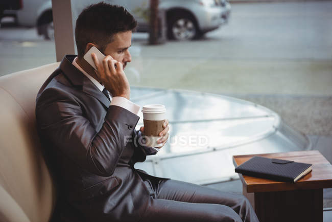 Homme d'affaires tenant un café tout en parlant sur un téléphone portable dans les locaux du bureau — Photo de stock