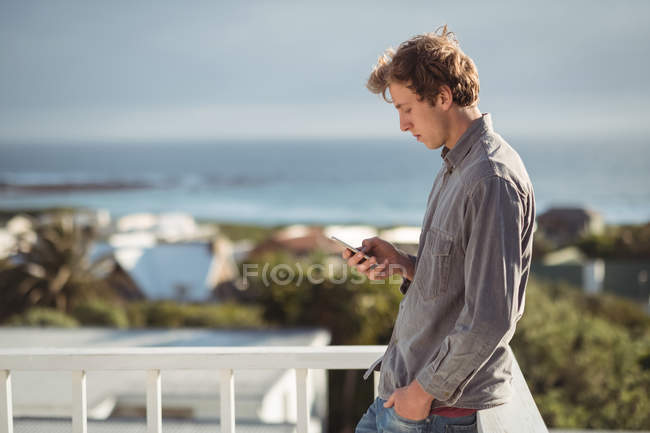 Vista lateral de un hombre apoyado en barandillas de balcón y usando teléfono móvil - foto de stock