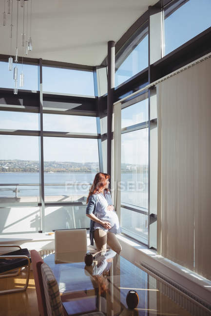 Femme enceinte réfléchie regardant par la fenêtre dans le salon à la maison — Photo de stock