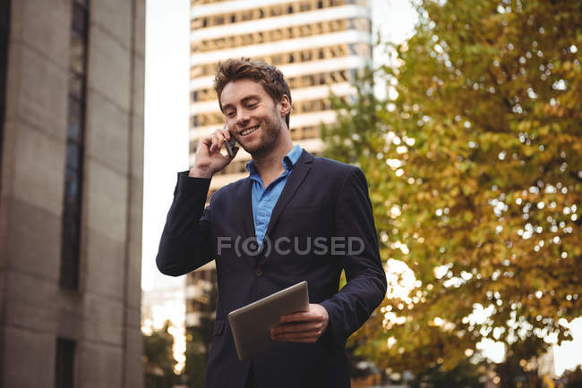 Lächelnder Geschäftsmann, der mit dem Handy telefoniert und auf der Straße ein digitales Tablet hält — Stockfoto