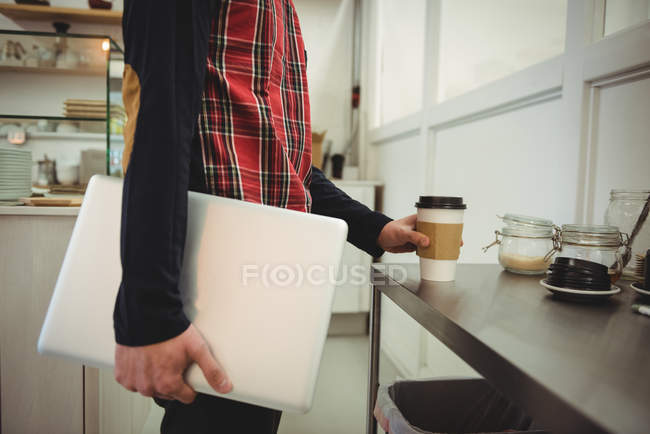 Mid-section of man holding a laptop and disposable coffee cup in coffee shop — Stock Photo