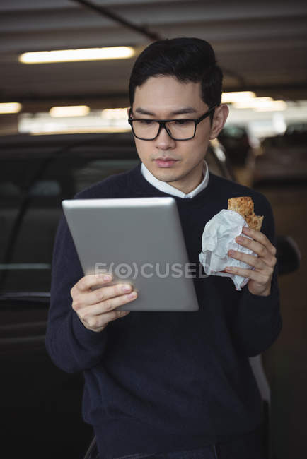 Businessman using digital tablet while eating snack in garage — Stock Photo
