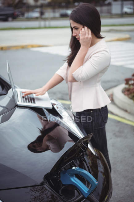 Mulher bonita falando no telefone celular enquanto carrega carro elétrico na rua — Fotografia de Stock