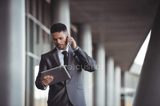 Businessman using digital tablet while talking on mobile phone in the office campus — Stock Photo