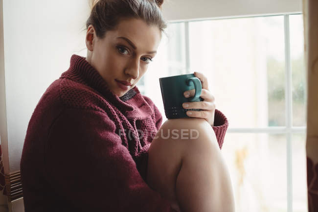 Retrato de mulher sentada na soleira da janela e segurando xícara de café em casa — Fotografia de Stock