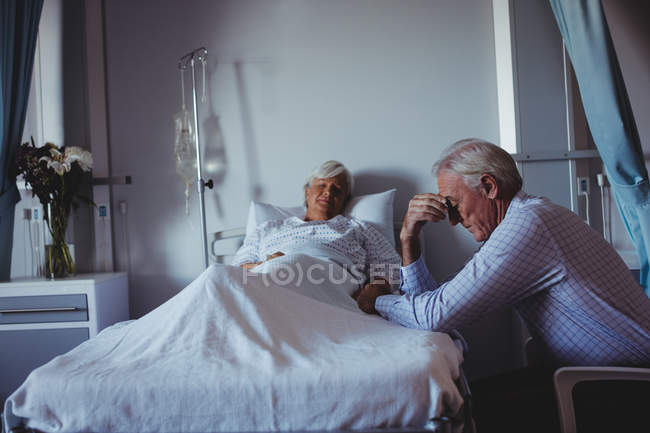 Mujer enferma durmiendo en la cama mientras hombre preocupado sentado al lado de su cama en el hospital - foto de stock