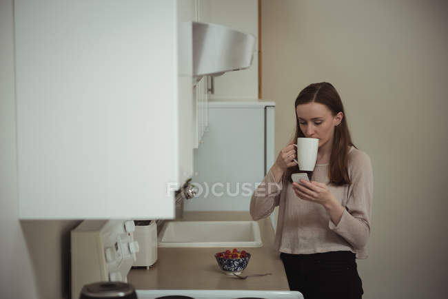 Жінка дивиться на мобільний телефон, маючи каву на кухні вдома — стокове фото