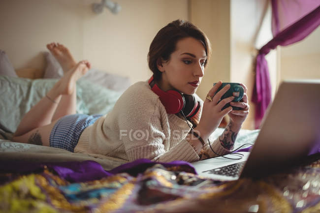 Beautiful woman using laptop while having coffee on bed at home — Stock Photo
