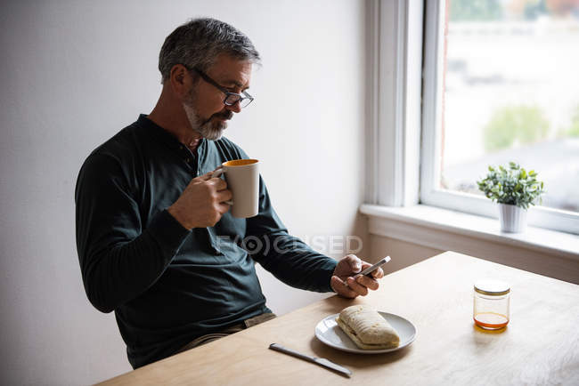 Homem usando telefone celular enquanto toma uma xícara de café em casa — Fotografia de Stock