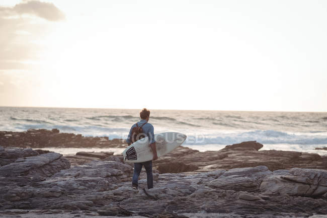 Hombre llevando una tabla de surf caminando hacia el mar al atardecer - foto de stock