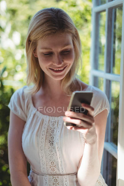 Mulher bonita sentada na janela e usando telefone celular em casa — Fotografia de Stock