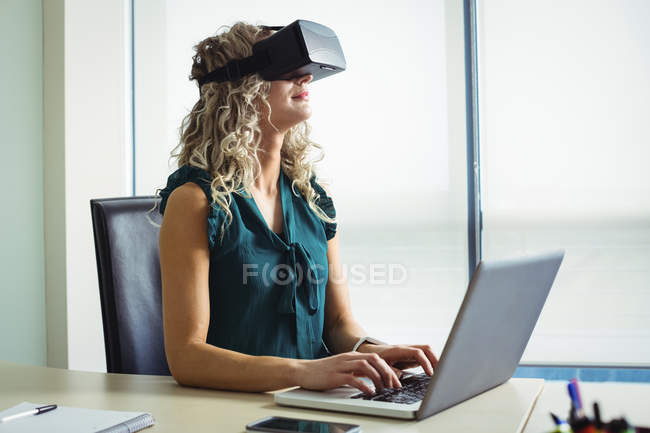 Business executive using virtual reality headset and working on laptop in office — Stock Photo