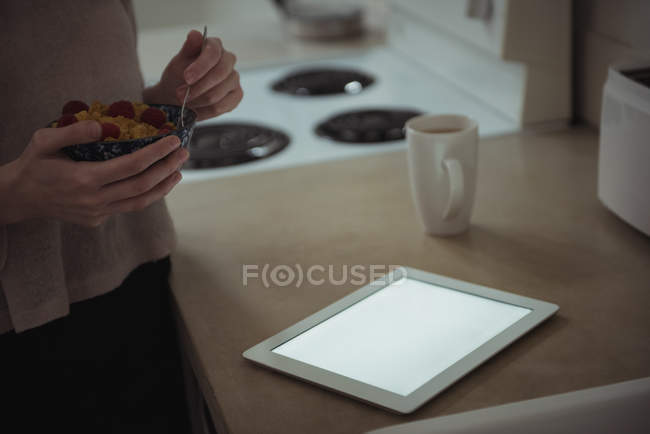 Sección media de la mujer con tazón de desayuno de pie cerca de la tableta digital en casa - foto de stock