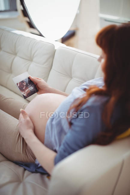 Pregnant woman relaxing on sofa and looking at sonography in living room — Stock Photo